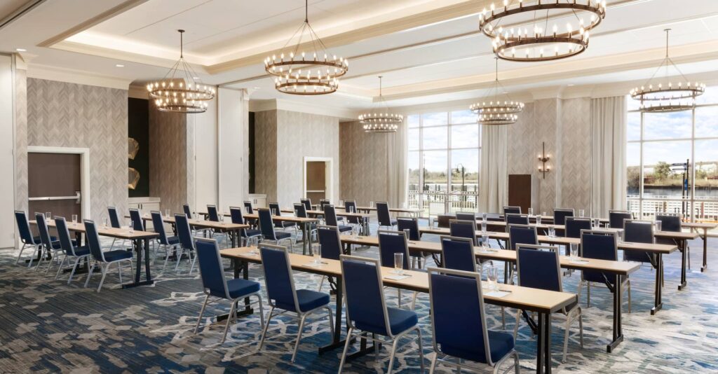 A bright and modern conference room with rows of tables and chairs, elegant chandeliers, and large windows offering views of the outdoors during Embassy Suites Breakfast Hours.