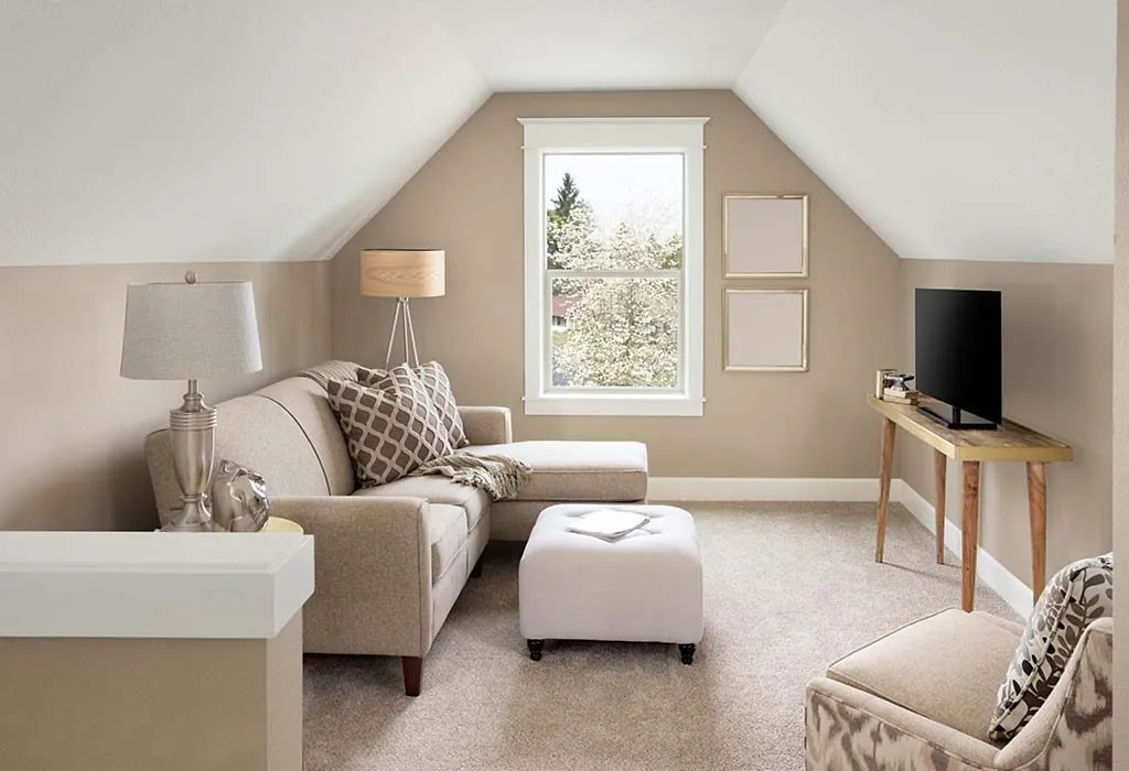 An attic living room with a beige sofa, patterned pillows, and a white ottoman. A wooden TV stand with a flat-screen TV is against the wall. Two empty frames hang on the wall, and a window offers a view of trees. A floor lamp is in the corner.