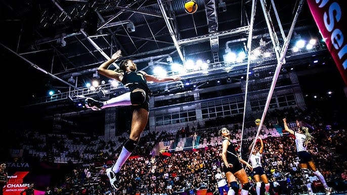 A volleyball player jumps high near the net, poised to spike the ball, with a packed stadium in the background. Teammates and opponents watch the action, and a vibrant arena atmosphere is evident with bright lights overhead.