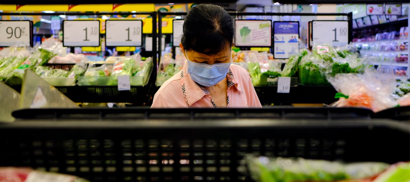 A woman wearing a face mask is shopping in a supermarket's produce section. She is surrounded by neatly arranged vegetables in plastic bags, with bright overhead lighting illuminating the scene.