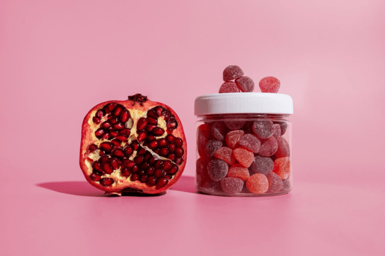 A halved pomegranate sits next to a clear jar filled with red and pink gummy candies against a pink background.